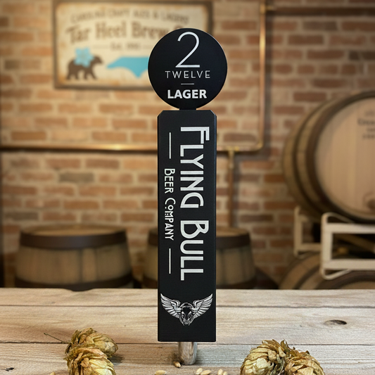 Flying Bull Beer Company tap handle with hops on a wooden surface against a brick wall.